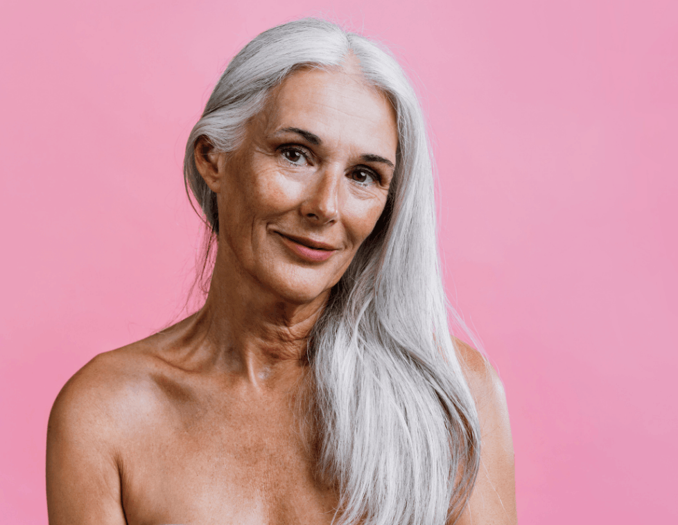 Smiling mature woman with long gray hair against a pink background.