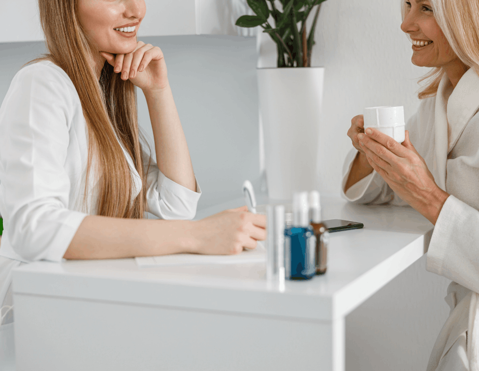 Two women chatting over coffee at a white table with a plant in the background.