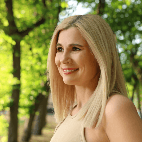 Smiling woman with blonde hair stands outdoors in a sunlit park.