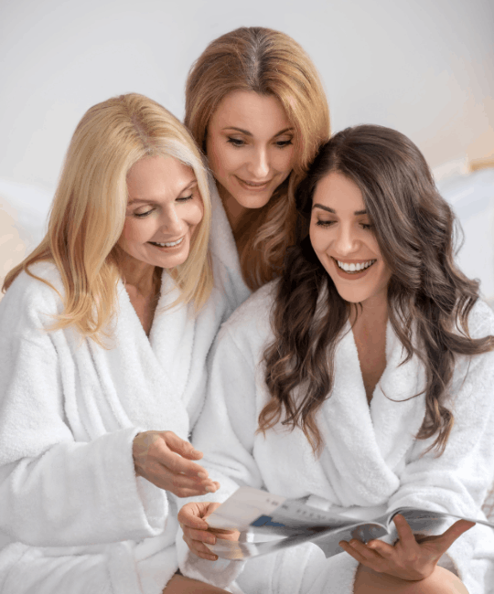 Three women in white robes looking at a phone and smiling.