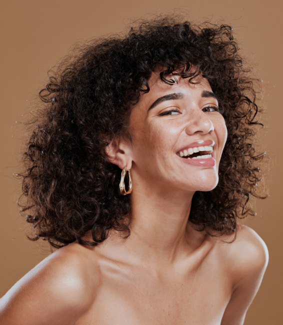 A woman with curly hair and hoop earrings smiles brightly against a neutral background.