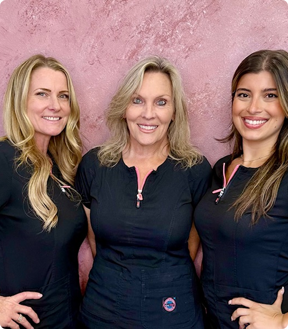 Three smiling women in black tops posing against a pink background.