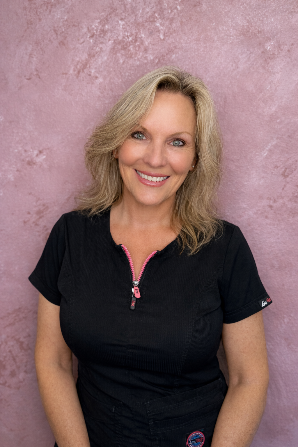 Smiling woman with blonde hair wearing a black top against a pink textured wall.